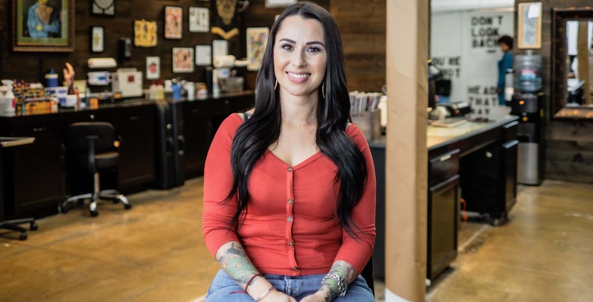 A smiling woman with long brown hair and arm tattoos wearing a red top and blue jeans sits in a workshop.