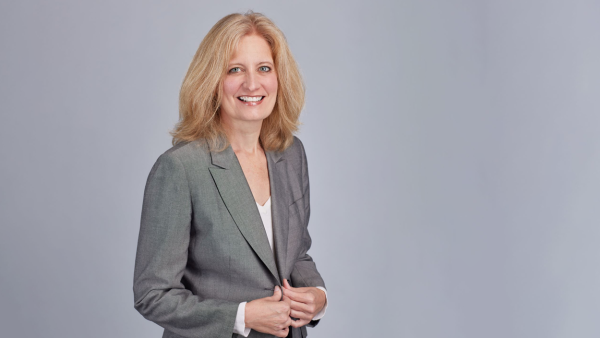 A blonde woman in a grey suit poses for a professional photo in front of a grey background.