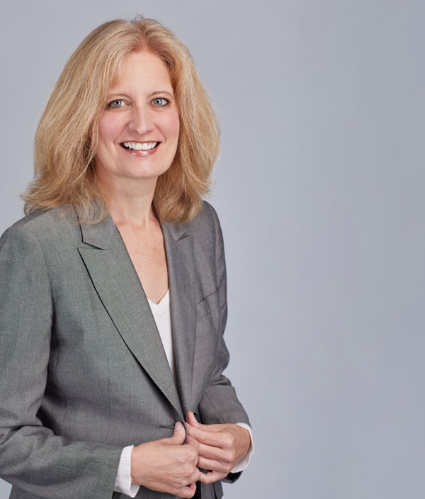 A blonde woman in a grey suit poses for a professional photo in front of a grey background.