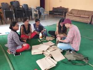 Five students sit on a green carpet with books and papers strewn in the middle.