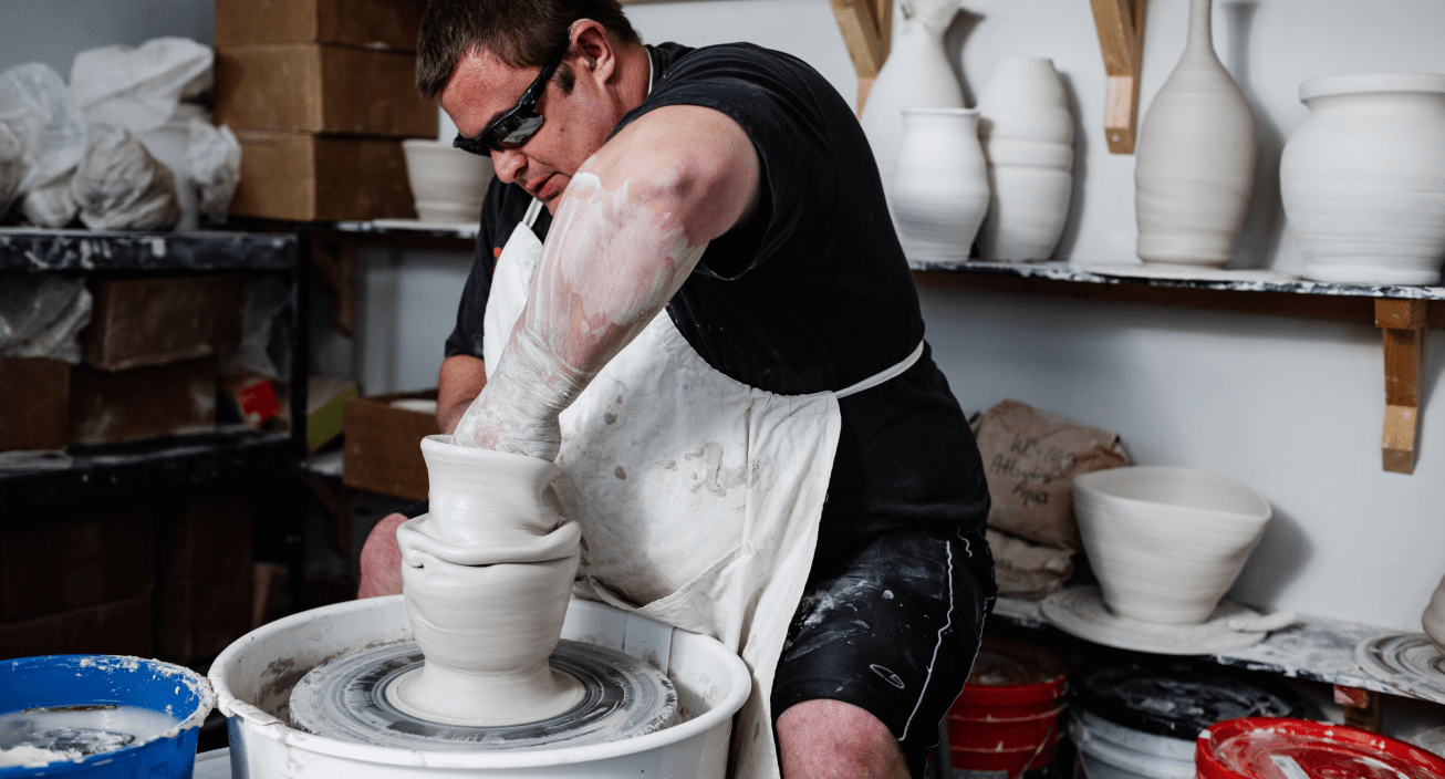 A man wearing sunglasses and a white apron molds clay on a potters wheel with finished vases and bowls in the background.