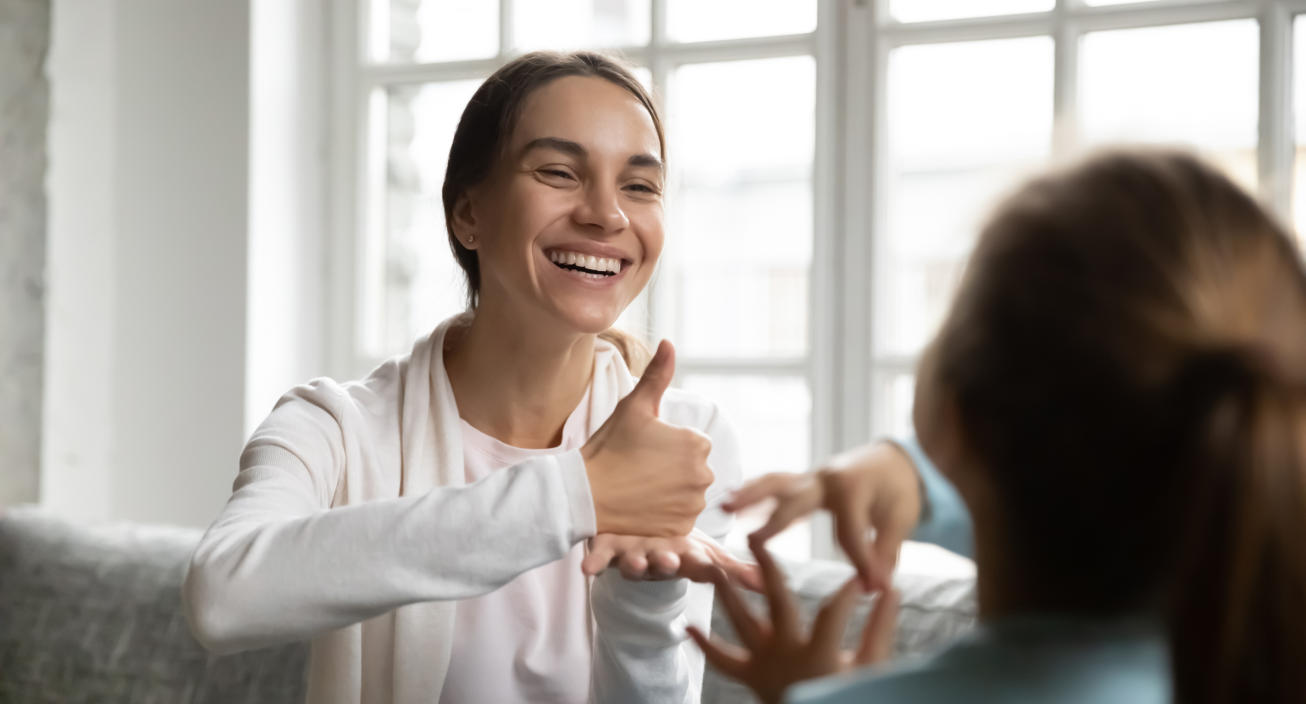 A smiling female wearing a white hoodie uses ASL to communicate.