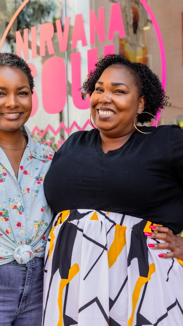 Two smiling Black women pose in front of their coffee shop with a pink window decal promoting Nirvana Soul.