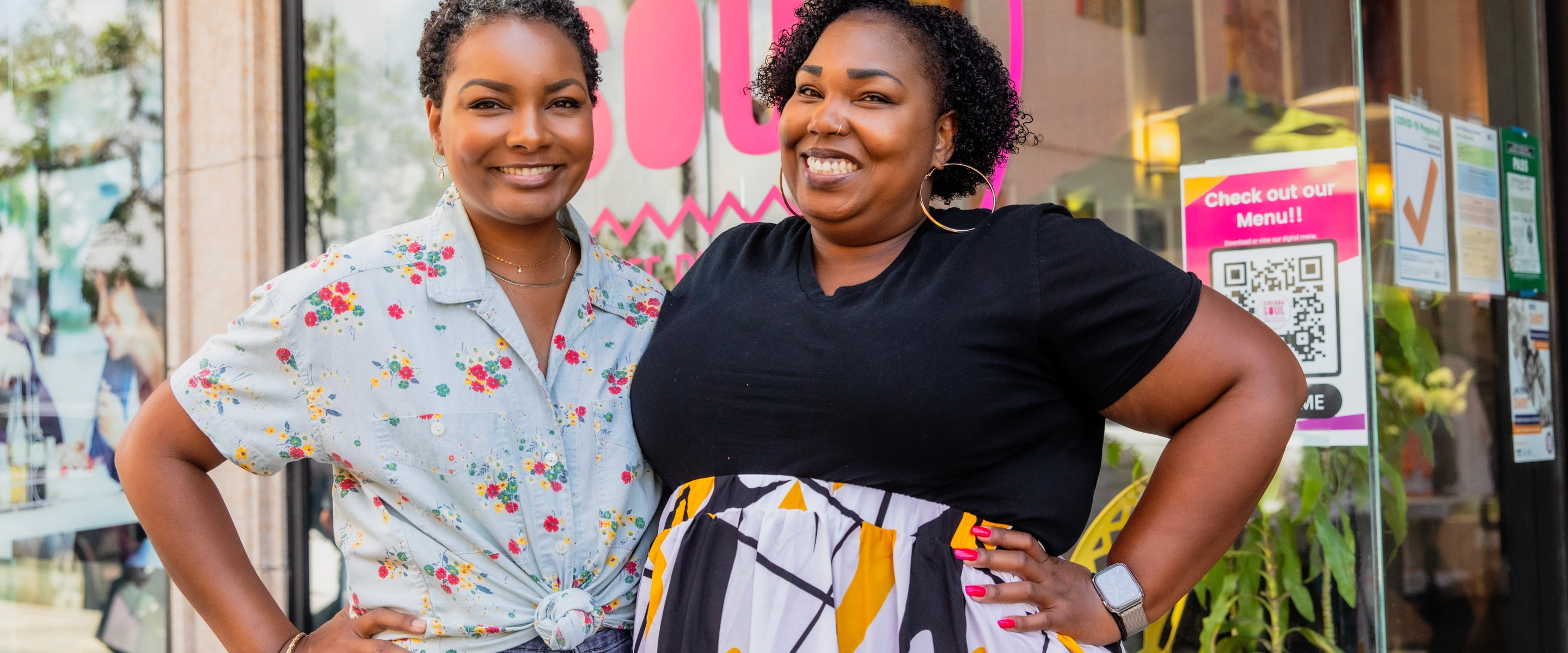 Two smiling Black women pose in front of their coffee shop with a pink window decal promoting Nirvana Soul.