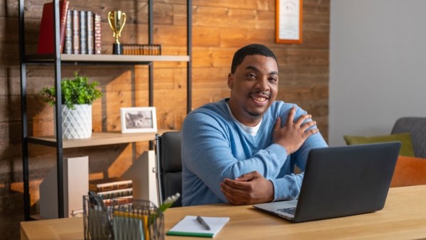 A man in a blue sweather smiles while working at his open laptop in a nicely adorned, masculine-looking office.