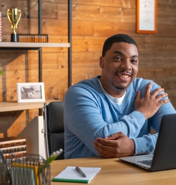 A man in a blue sweather smiles while working at his open laptop in a nicely adorned, masculine-looking office.