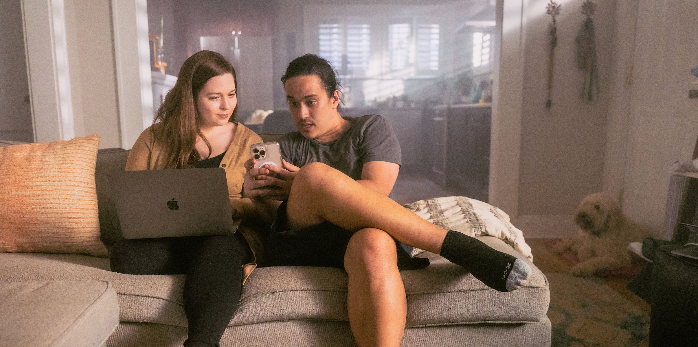 A woman and man sit on the couch with an open laptop while looking a a phone.