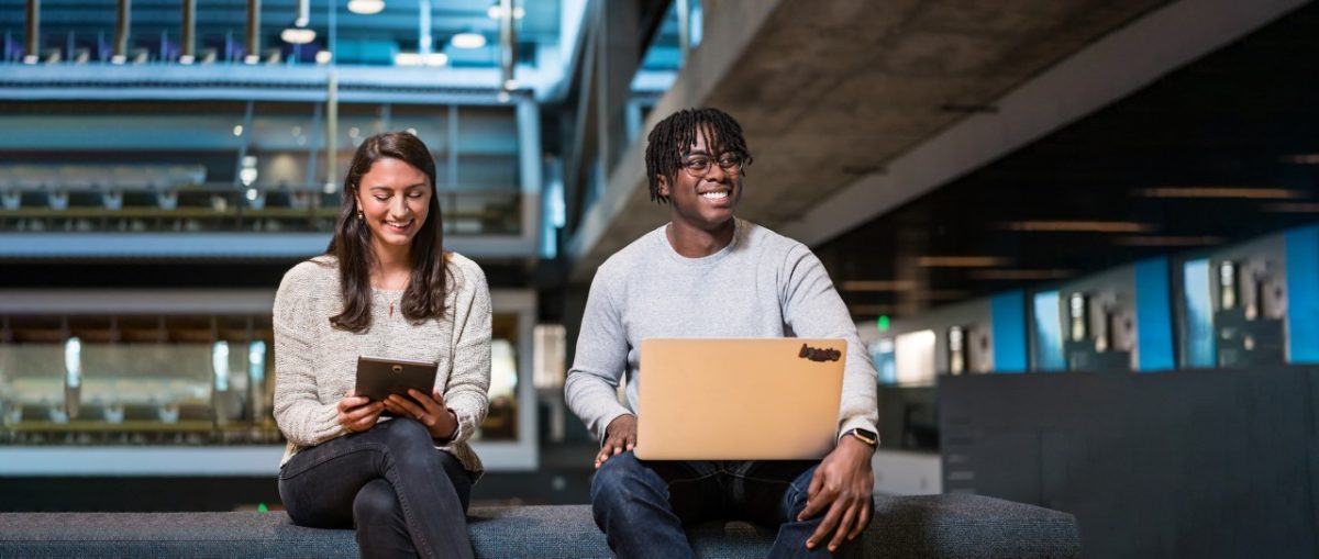 A brunette tech worker in her 20s smiles while looking at her tablet in an industrial chic open office setting. Next to her a young, Black man with short braids works at his computer.