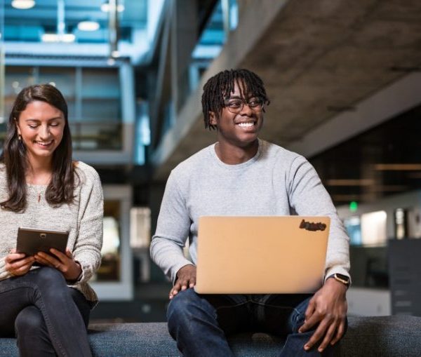 A brunette tech worker in her 20s smiles while looking at her tablet in an industrial chic open office setting. Next to her a young, Black man with short braids works at his computer.