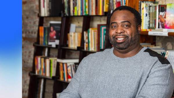 A professional headshot of a Black man in a grey sweater, smiling at the camera with a colorful bookcase in the background.