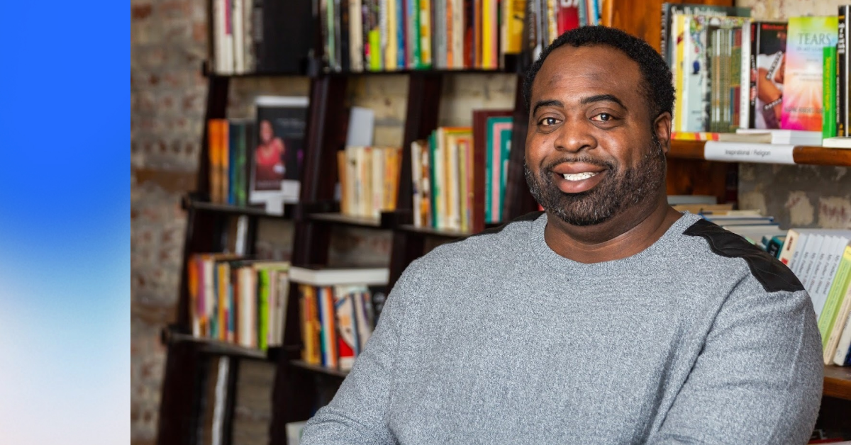 A professional headshot of a Black man in a grey sweater, smiling at the camera with a colorful bookcase in the background.