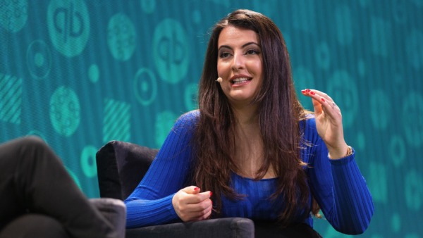 A brunette woman in a blue sweather speaks at a conference wearing a headset mic.