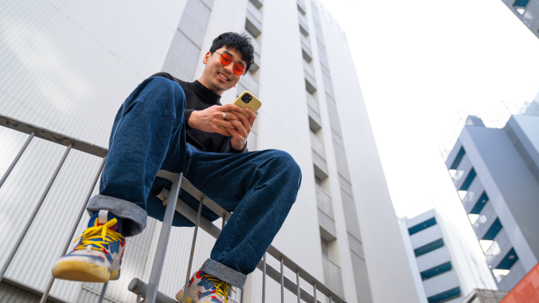 A young man sits on a fence in a city, looking at his phone.