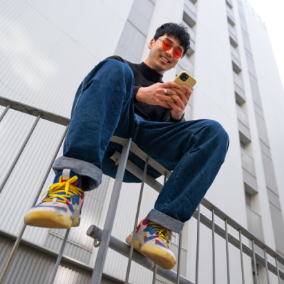 A young man sits on a fence in a city, looking at his phone.