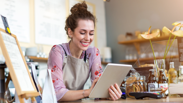 A woman with curly, red hair uses her tablet while wearing an apron and leaning over a counter at ther business.