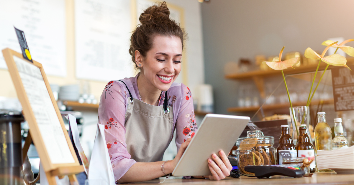 A woman with curly, red hair uses her tablet while wearing an apron and leaning over a counter at ther business.