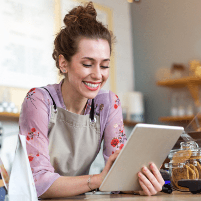 A woman with curly, red hair uses her tablet while wearing an apron and leaning over a counter at ther business.