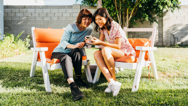 An Asian mother and daughter sit outside under an umbrella, looking at a phone and smiling.